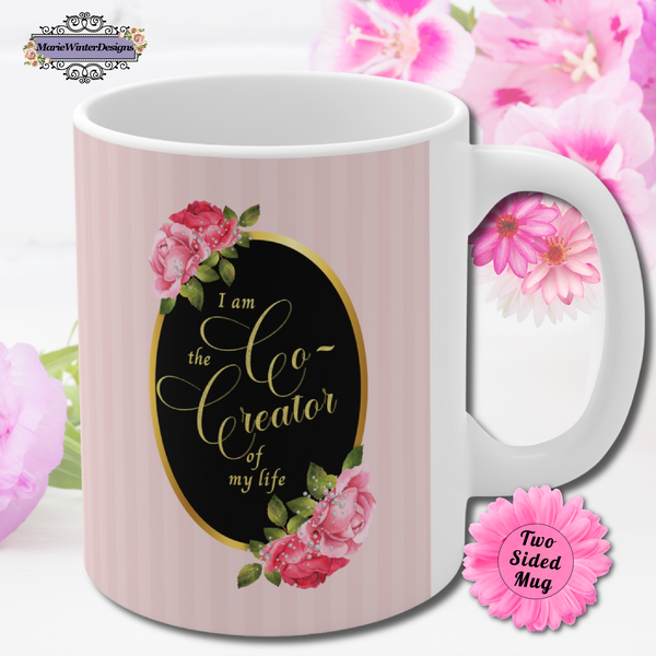 ceramic mug with saying" I am the co-creator of my life" written in gold lettering on a black oval with gold edge accented with large pink roses at the top and bottom of the oval on a background of pink stripes. Pink flowers behind the mug.