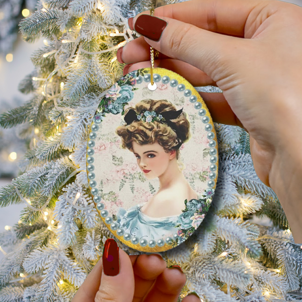 Hands with dark red nail polish holding an oval Ceramic Ornament With Elegant Early 1900s Vintage Harrison Fisher Illustration of Lady with Blue Flowers Surrounded with Blue Pearls with Christmas Tree branches in background