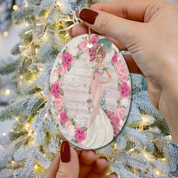 Hands with dark red nail polish holding an oval Oval ceramic ornament with Edwardian Lady with large hat music sheet background surrounded with pink and white roses with Christmas tree in background.