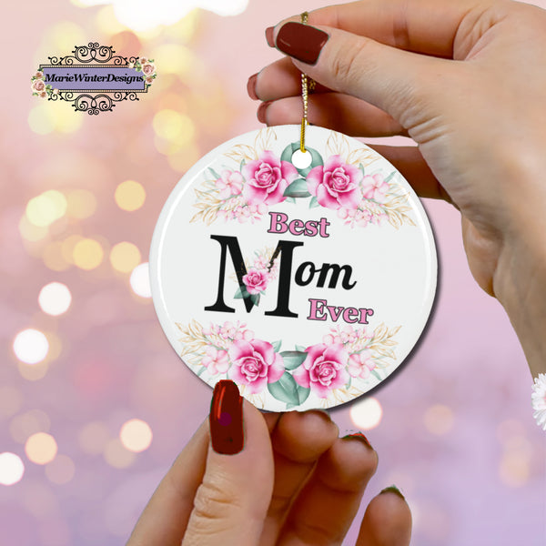 A person with red nails holds a "Best Mom Ever" floral ornament featuring pink roses and greenery, hanging by a gold string. The background is a soft bokeh effect in shades of pink and gold.