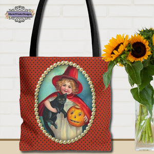 A Halloween tote bag featuring a vintage-style illustration of a young girl in a red witch costume holding a black cat and a jack-o-lantern, framed by pearls on a polka-dot orange background. The bag is placed next to a vase of vibrant sunflowers