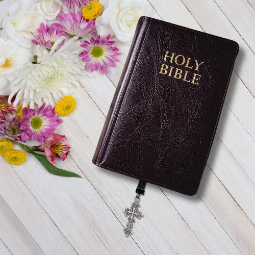 A dark brown textured Holy Bible with gold embossed lettering, shown closed on a white wood plank background with decorative pink, white, and yellow flowers and a silver cross bookmark.