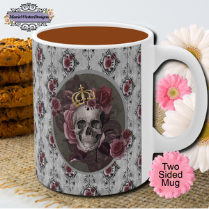 Ceramic mug with a skull wearing a gold crown accented with burgundy roses inside an oval with a light gray border, on a background of gray with black scrolls and burgundy roses with white handle. Pink and white flowers along with a stack of cookies are behind the mug.