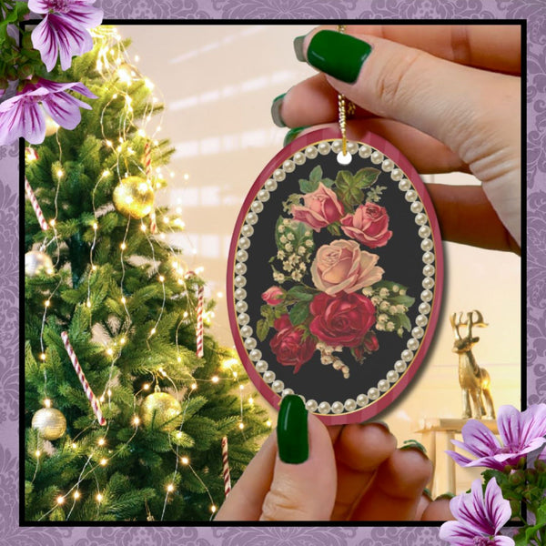 hands with green nail polish holding an oval ceramic ornament Vintage Floral Cameo Design with red and pink flowers against black background surrounded with a fame of cream pearls with a Christmas tree in the background of room