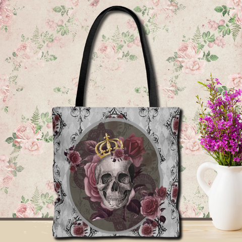 Tote bag with black handle and design of  a gray skull with gold crown and roses on a gray background with back scroll lines and burgundy flowers. 
