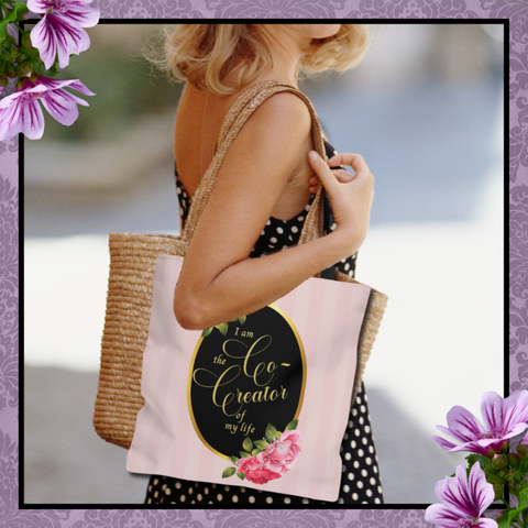 Woman wearing a black and white polka dot dress carrying a Tote bag that says "I am the co-creator of my life" written in gold letters on a black oval frame in gold with Pink roses on the top and bottom on a back ground of light pink stripes.