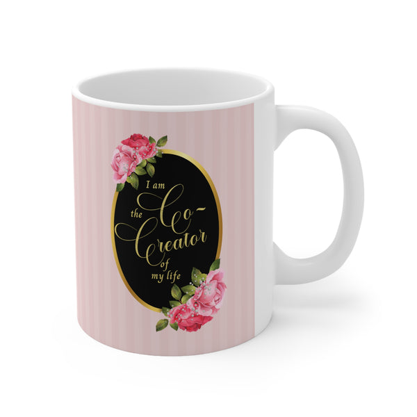 ceramic mug with saying" I am the co-creator of my life" written in gold lettering on a black oval with gold edge accented with large pink roses at the top and bottom of the oval on a background of pink stripes.