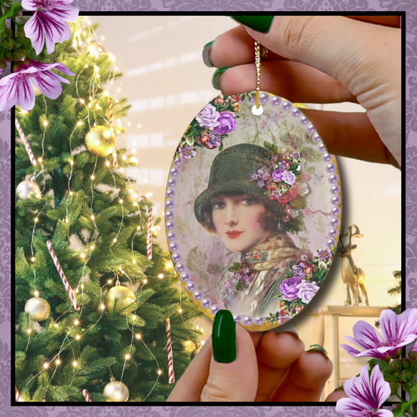 Hands wit green nail polish holding an oval Ceramic Ornament With Elegant Early 1900s Vintage Woman in Green Flapper Style Gatsby Hat surrounded by purple pearls on gold background with pink roses to the side with Christmas tree in the back.