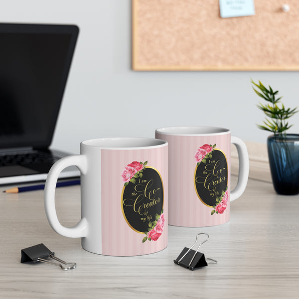 Front and back view of ceramic mug with saying" I am the co-creator of my life" written in gold lettering on a black oval with gold edge accented with large pink roses at the top and bottom of the oval on a background of pink stripes. Same both sides
